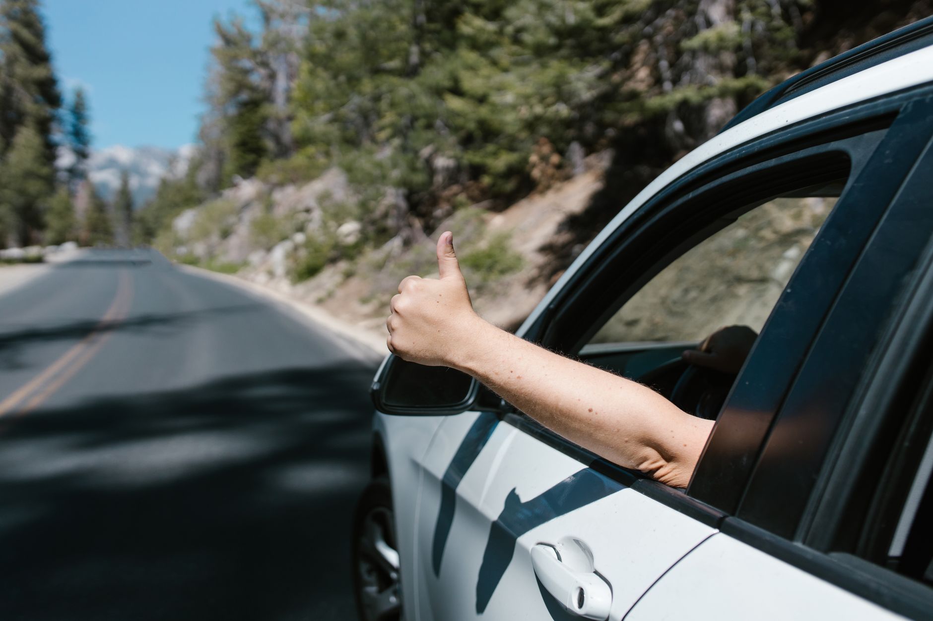 A person gives a thumbs up gesture from a car window while driving on a scenic forest road. — Safe Driving Suggestions To Fol